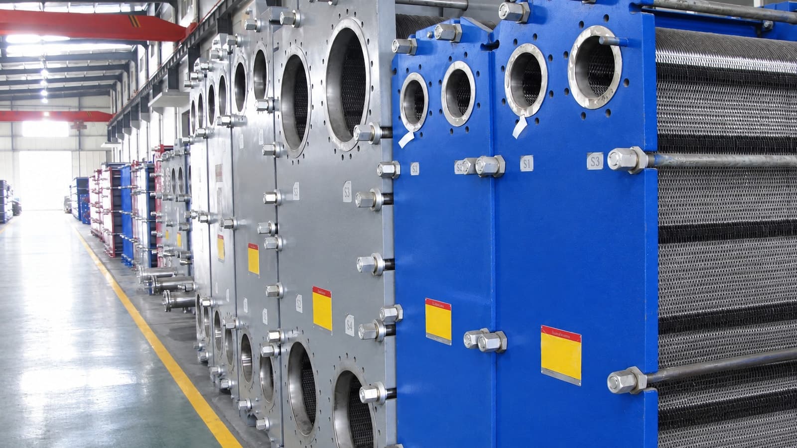 A line of finished gasketed plate heat exchangers down an assembly hall, ports flanged and tie bars tensioned
